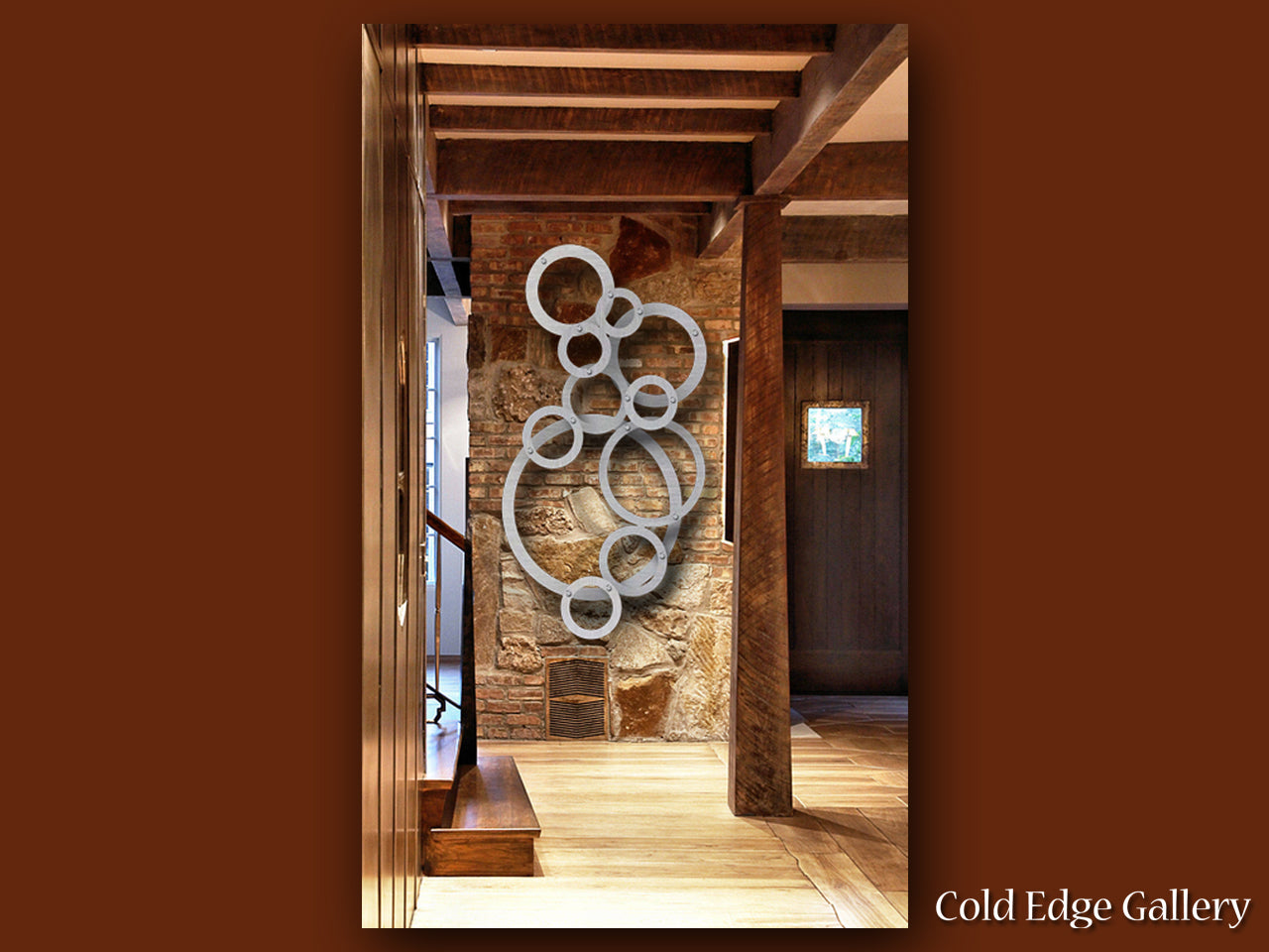 A custom wall-mounted metal sculpture of interlocking circles is displayed in a rustic interior with wooden beams and stone walls. The image includes the text "Cold Edge Gallery" in the bottom right corner.