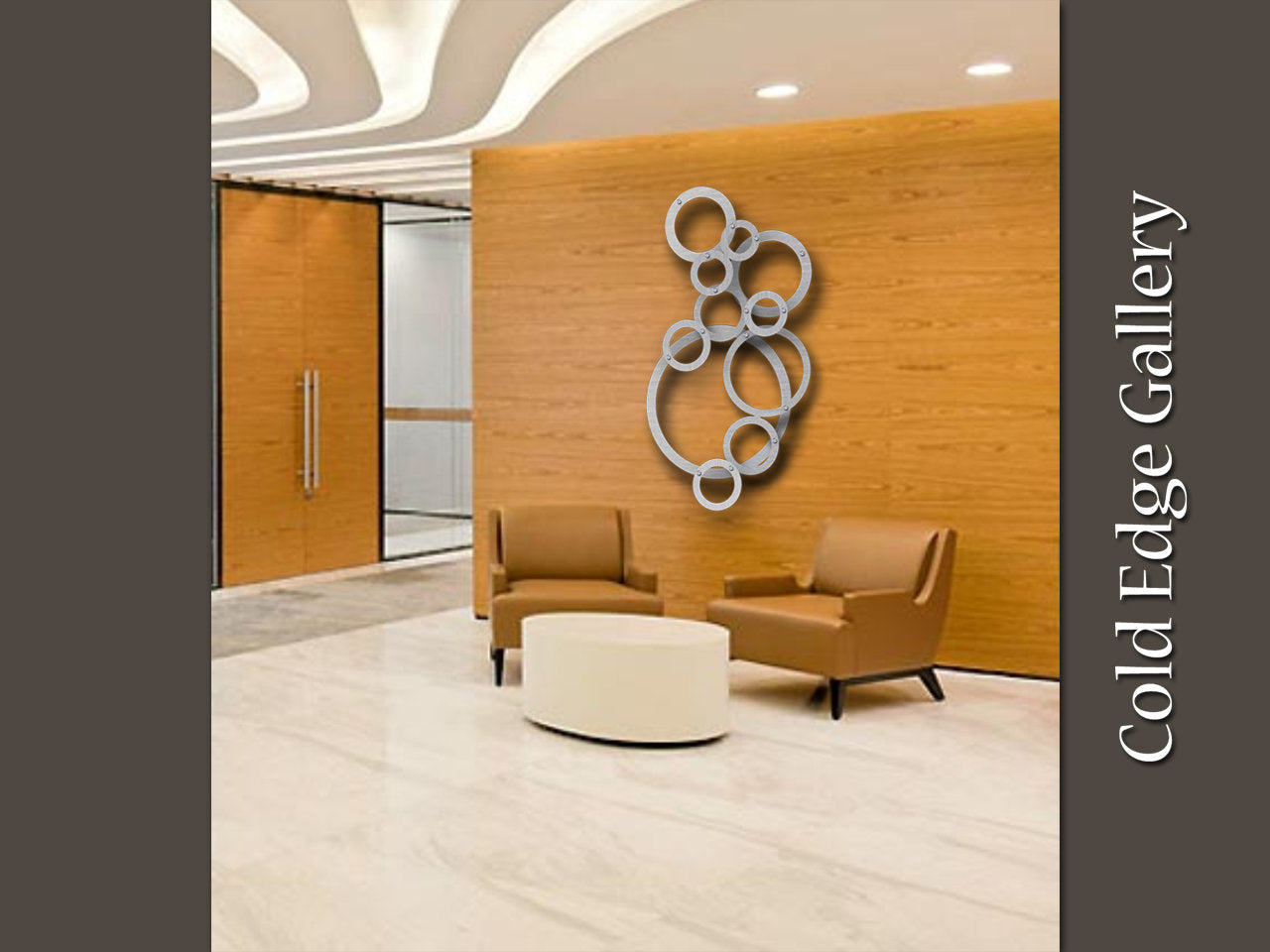 Lobby area with two brown leather chairs, a small round table, and a wooden accent wall featuring a circular custom metal artwork piece. A glass door allows light to stream in, and text on the wall reads "Cold Edge Gallery.