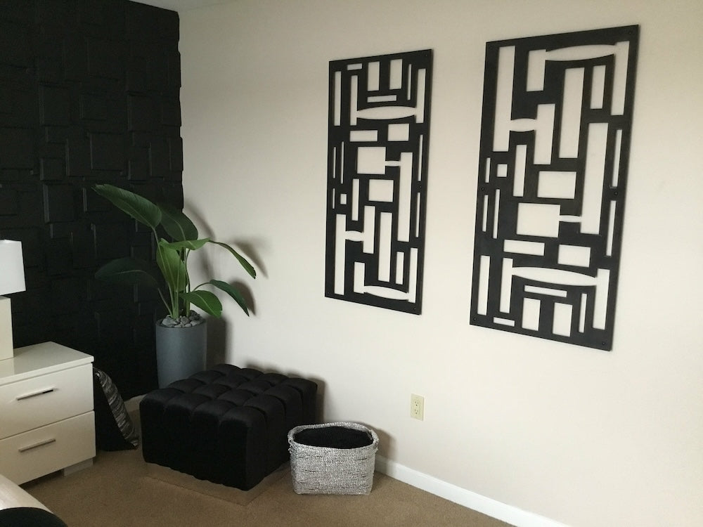 A room corner with two abstract black wall art pieces, a potted plant, a black ottoman, a woven basket, and a white dresser with a lamp.