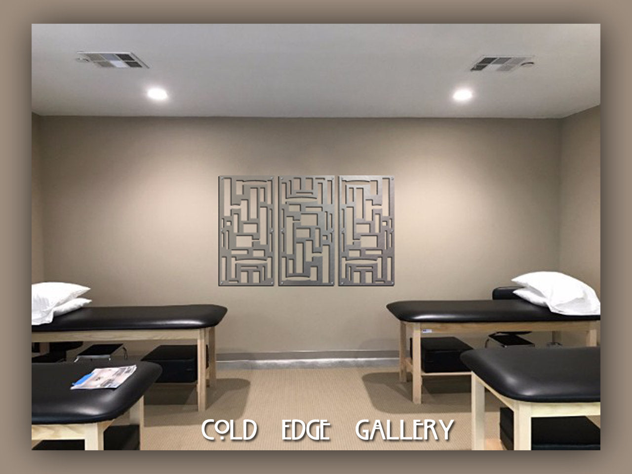 A room with two massage tables, each with a pillow. A geometrical metal wall art is centred on the back wall. The words "Cold Edge Gallery" are written at the bottom of the image.