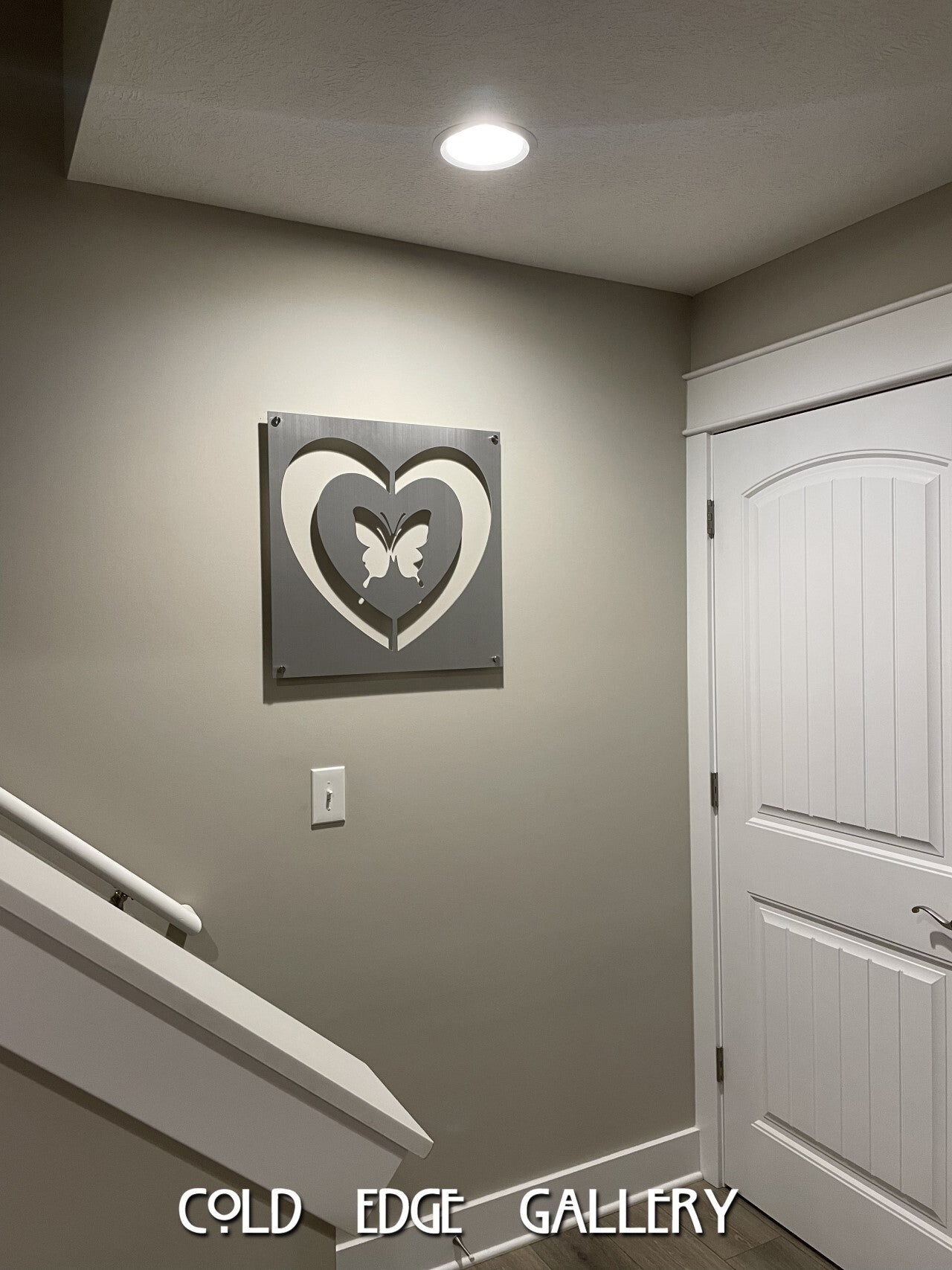 Framed art of two interlocking hearts with a butterfly in the center, mounted on a beige wall above a light switch near a white door. Cold Edge Gallery label at the bottom.