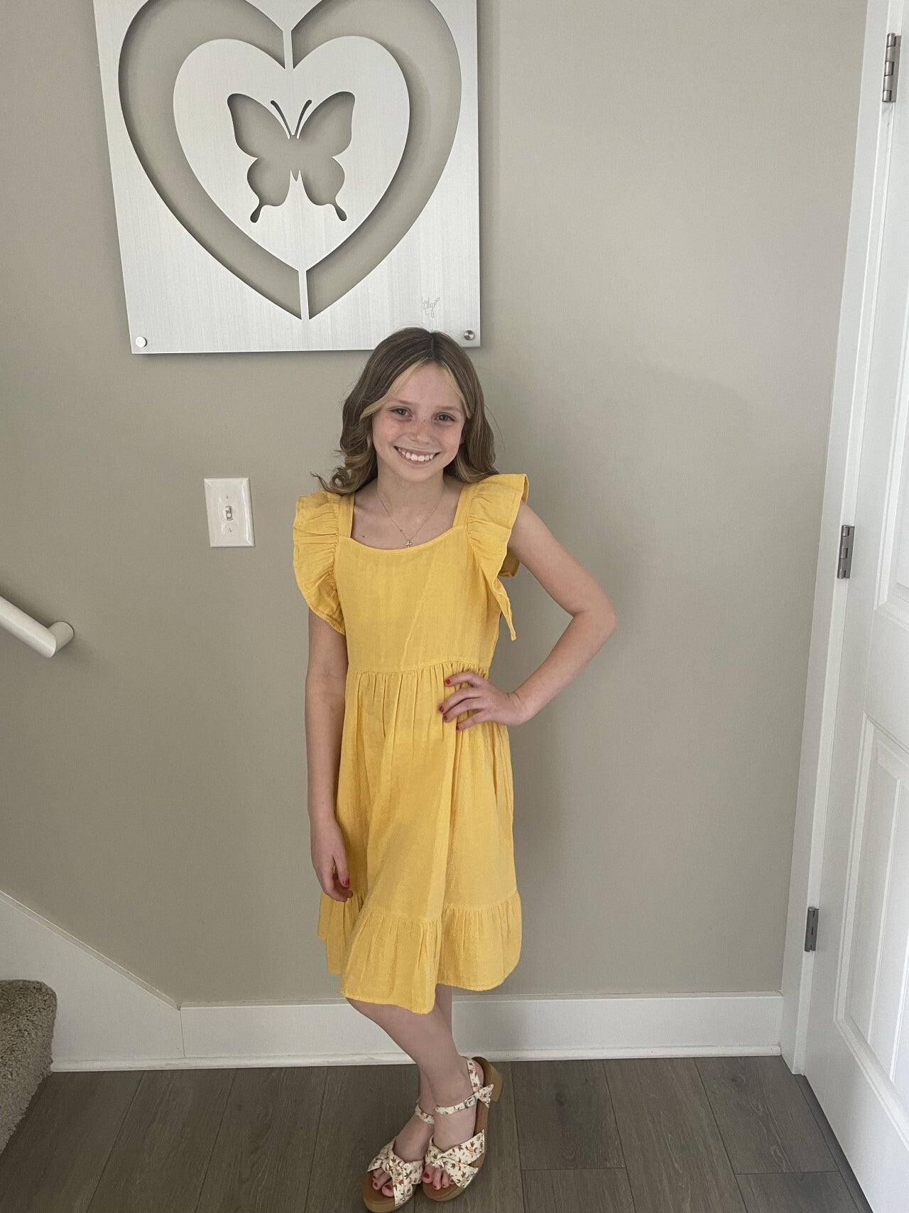 A young girl stands indoors, smiling, wearing a yellow dress with ruffled sleeves and brown high-heeled sandals. Behind her, there is a butterfly artwork in a heart frame on the wall.