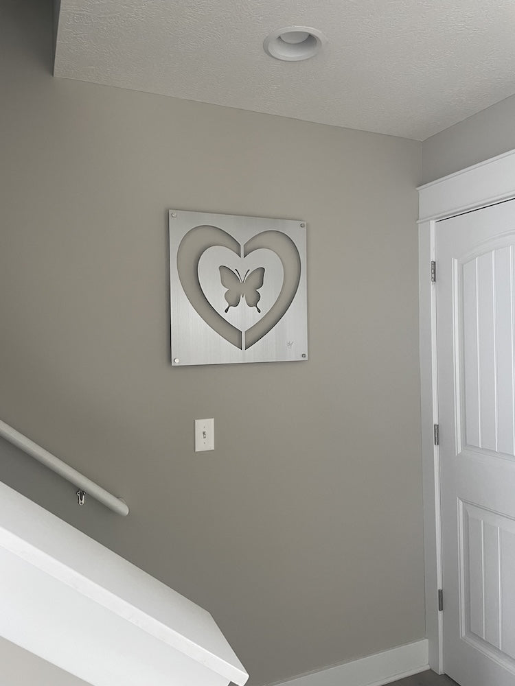 Wall art featuring a butterfly within a heart shape, mounted on a gray wall in a hallway near a white door and staircase.