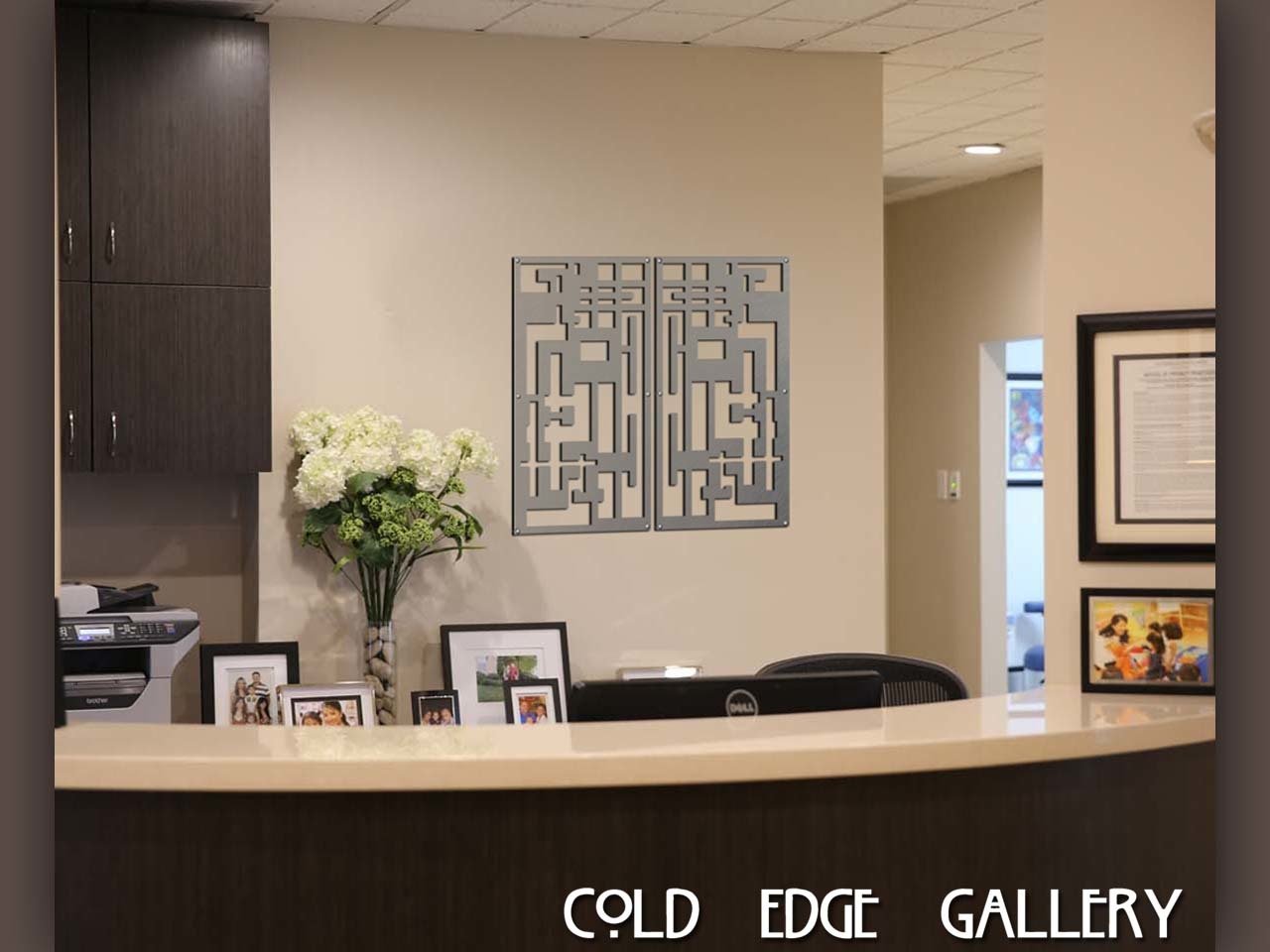 A reception desk with a curved counter, decorated with framed photos, flowers, and a modern geometric wall art piece behind. The text "COLD EDGE GALLERY" is displayed at the bottom.