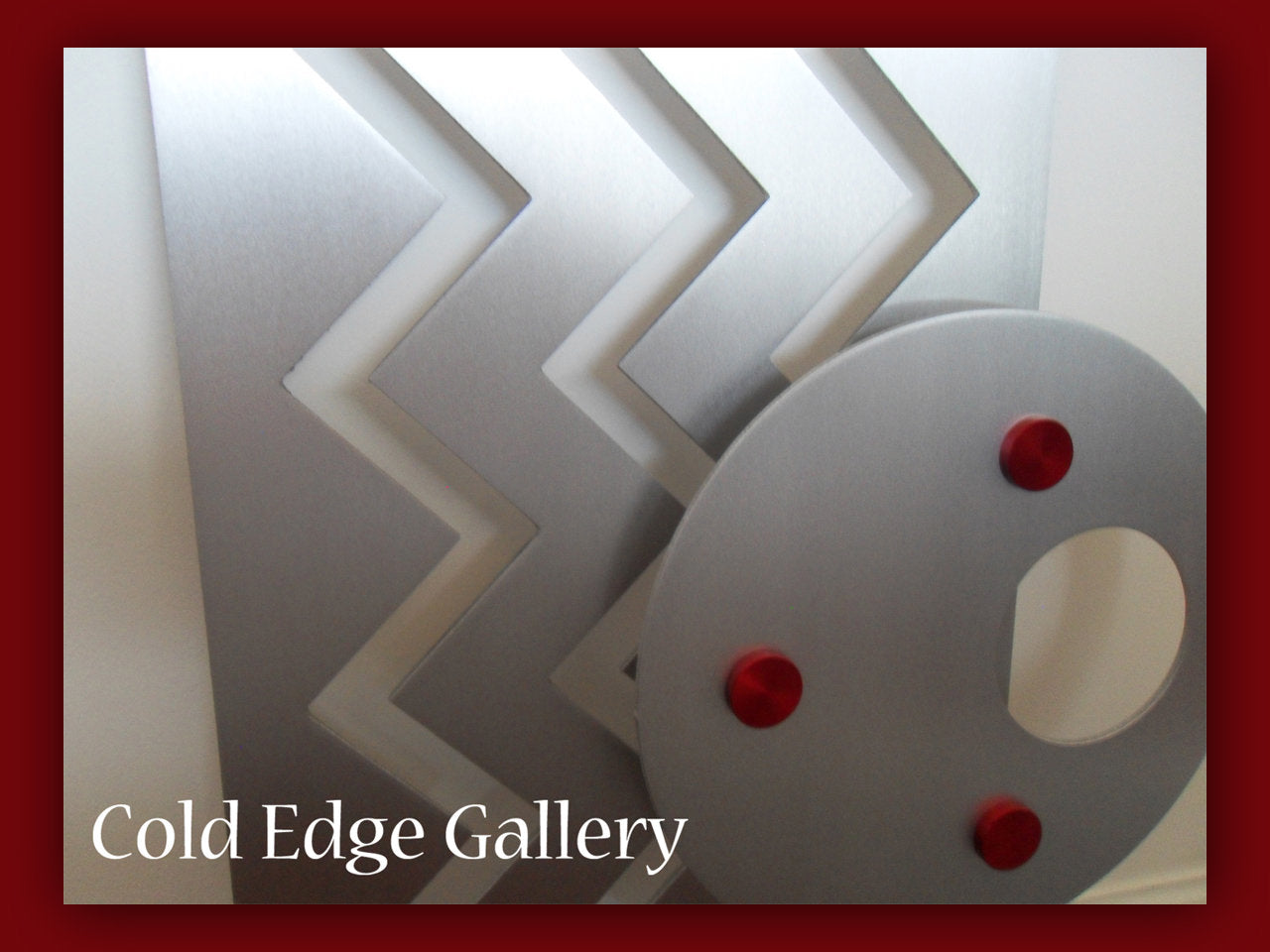 A geometric metal artwork featuring zigzag patterns and a circular element with red accents. The text "Cold Edge Gallery" is displayed in the lower-left corner.