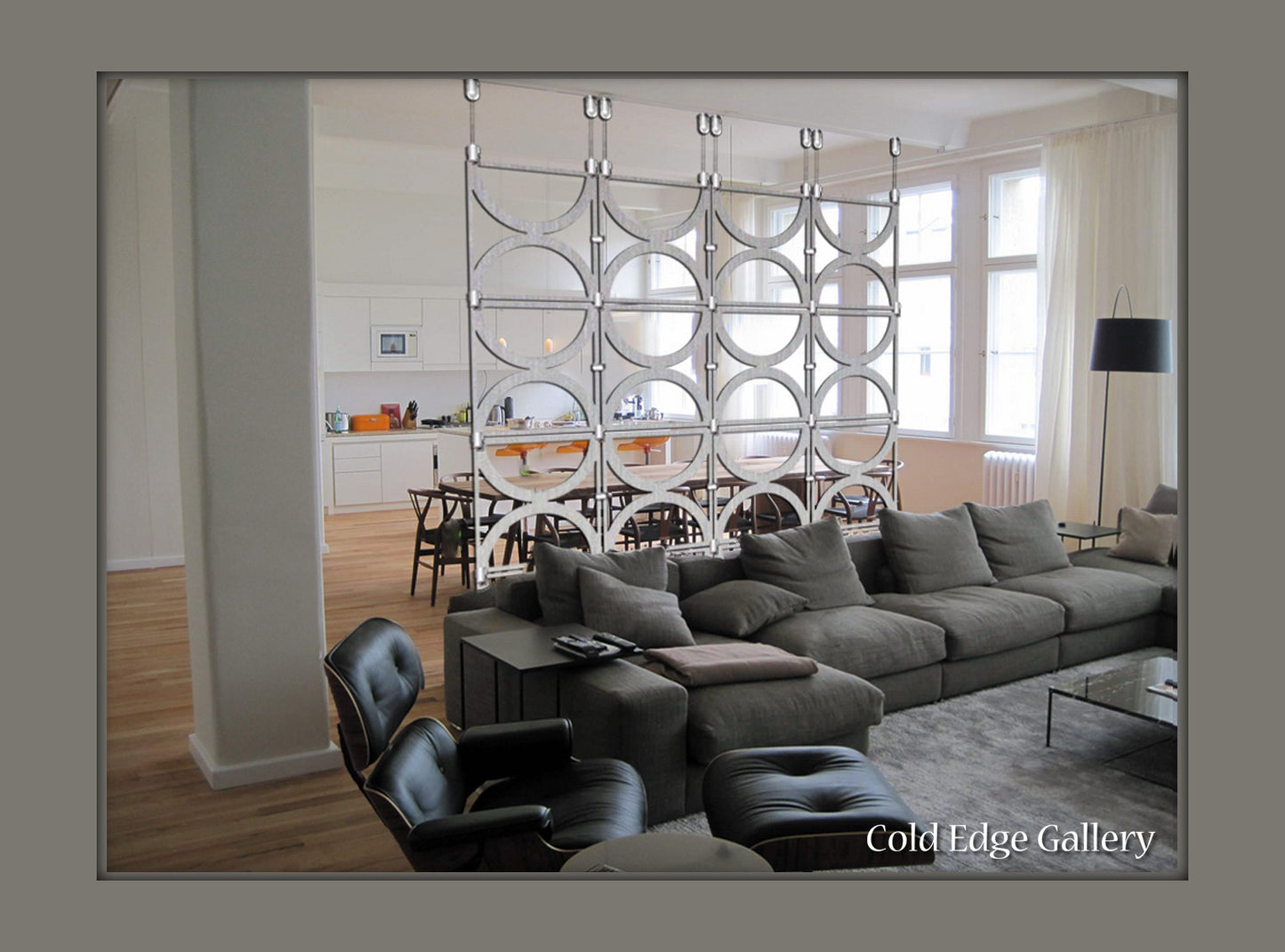 Modern living room with grey sectional sofa, lounge chair, and coffee table. Room divider with circular pattern separates living area from dining space and kitchen. Text reads "Cold Edge Gallery.