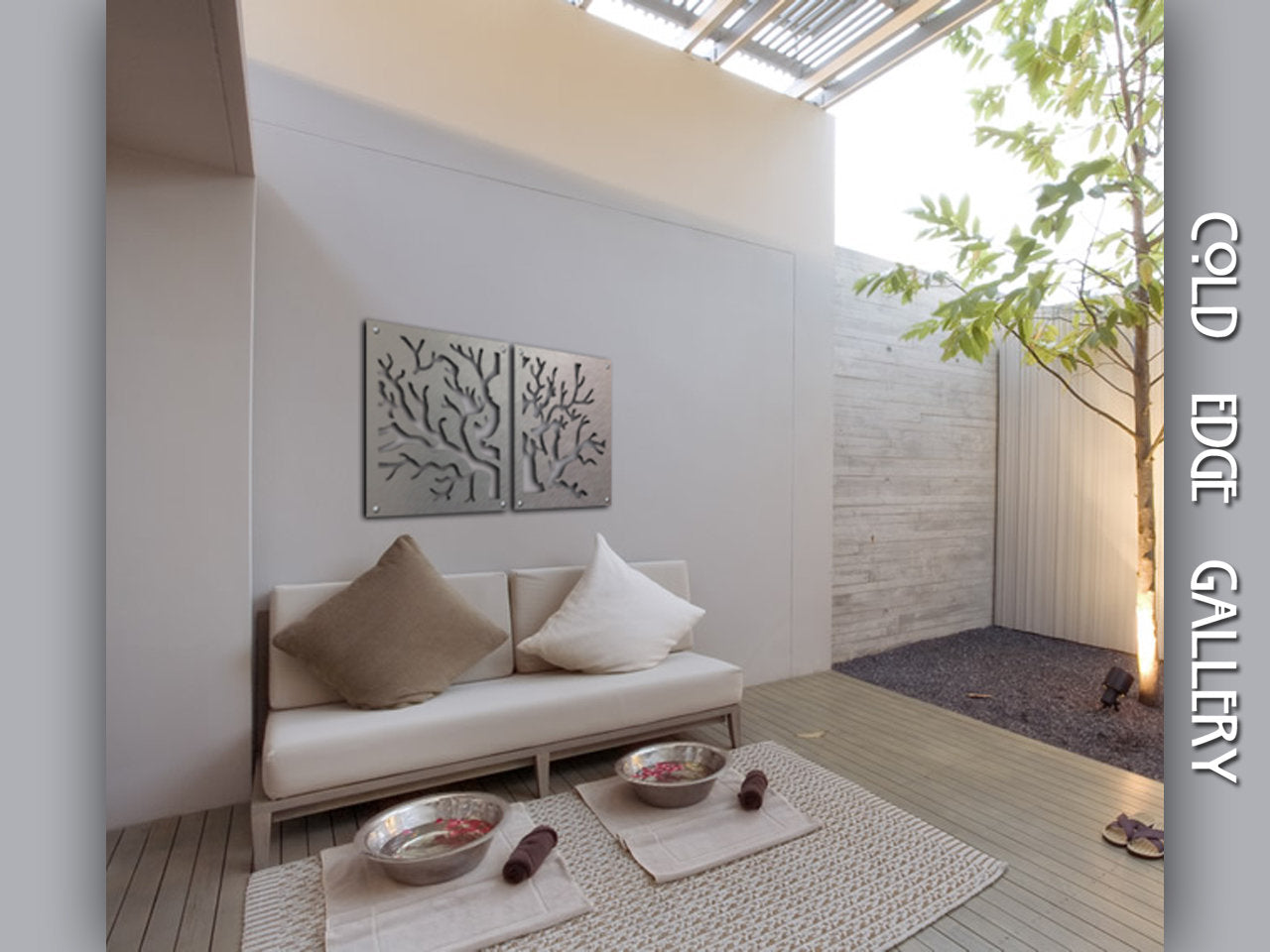 Outdoor minimalist seating area featuring a white couch with beige and brown pillows, metal tree art on the wall, and two metal bowls on a woven mat on the floor. A tree grows next to the space.