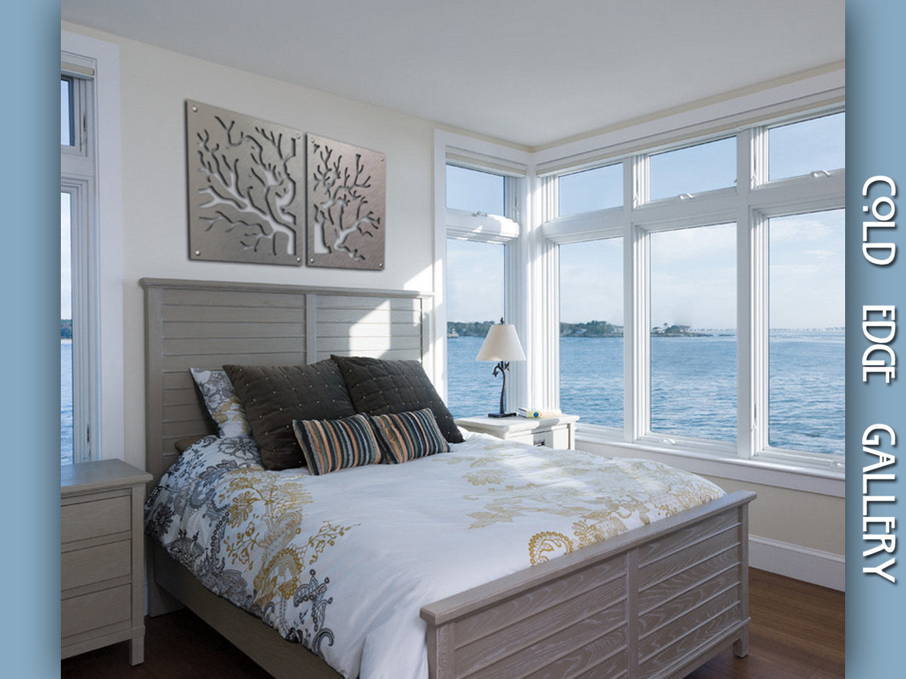 A well-lit bedroom with a large window overlooking a body of water. The bed has white bedding with floral patterns and multiple pillows. Two metal tree art pieces hang above the bed.