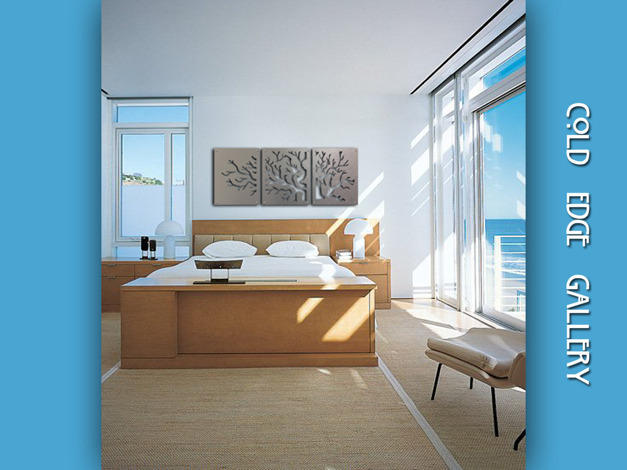 A modern bedroom featuring a light wood bed, matching furniture, and a triptych of tree-like metal artwork above the bed. The room has large windows with a view of the ocean. Text reads: "Cold Edge Gallery.