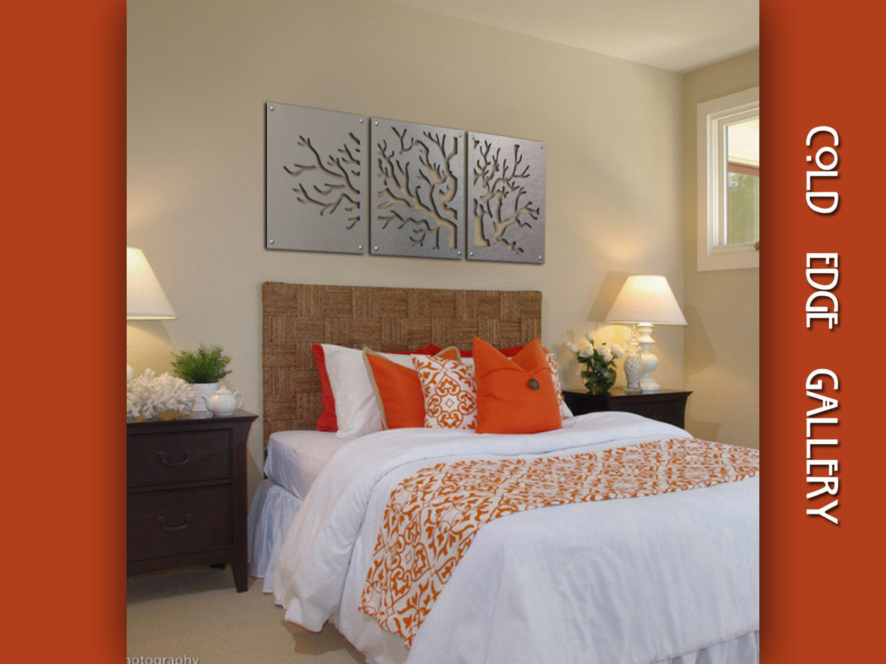 A bedroom with a wicker headboard features a bed with white and orange bedding. Above the bed is a three-piece metal wall art of trees. There are two nightstands with lamps on either side of the bed.