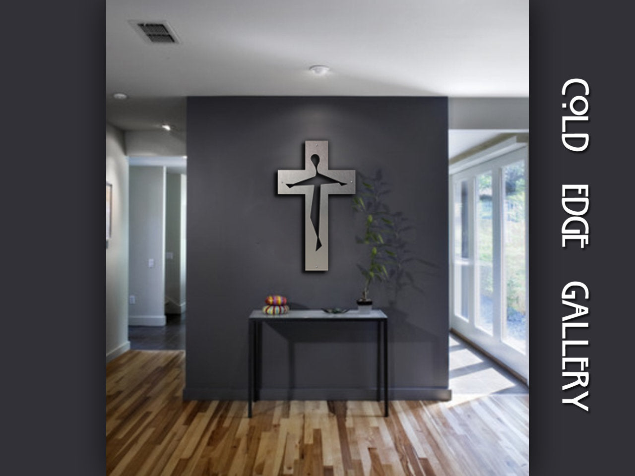 Art gallery interior with a modern cross sculpture on a dark grey wall, a slender black console table beneath it, and a plant. The walls have "Cold Edge Gallery" text vertically on the right side.