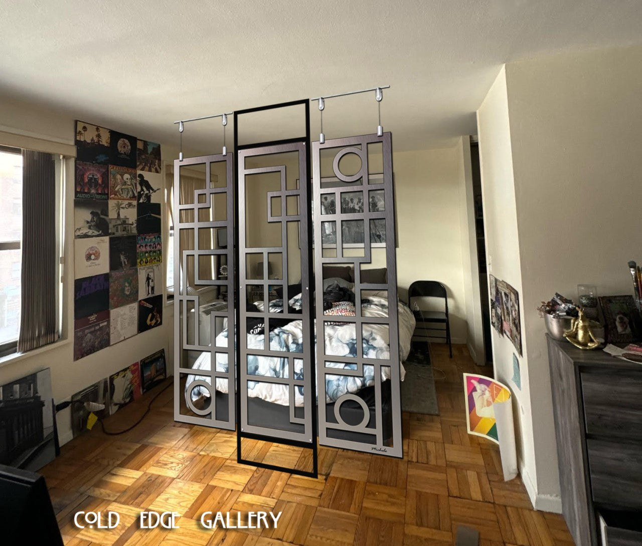 A room interior featuring a three-panel decorative metal divider with geometric patterns, partially separating a bedroom area. The panels hang from the ceiling, with the Cold Edge Gallery logo displayed at the bottom of the image. The room is decorated with posters on the walls and a wooden floor.