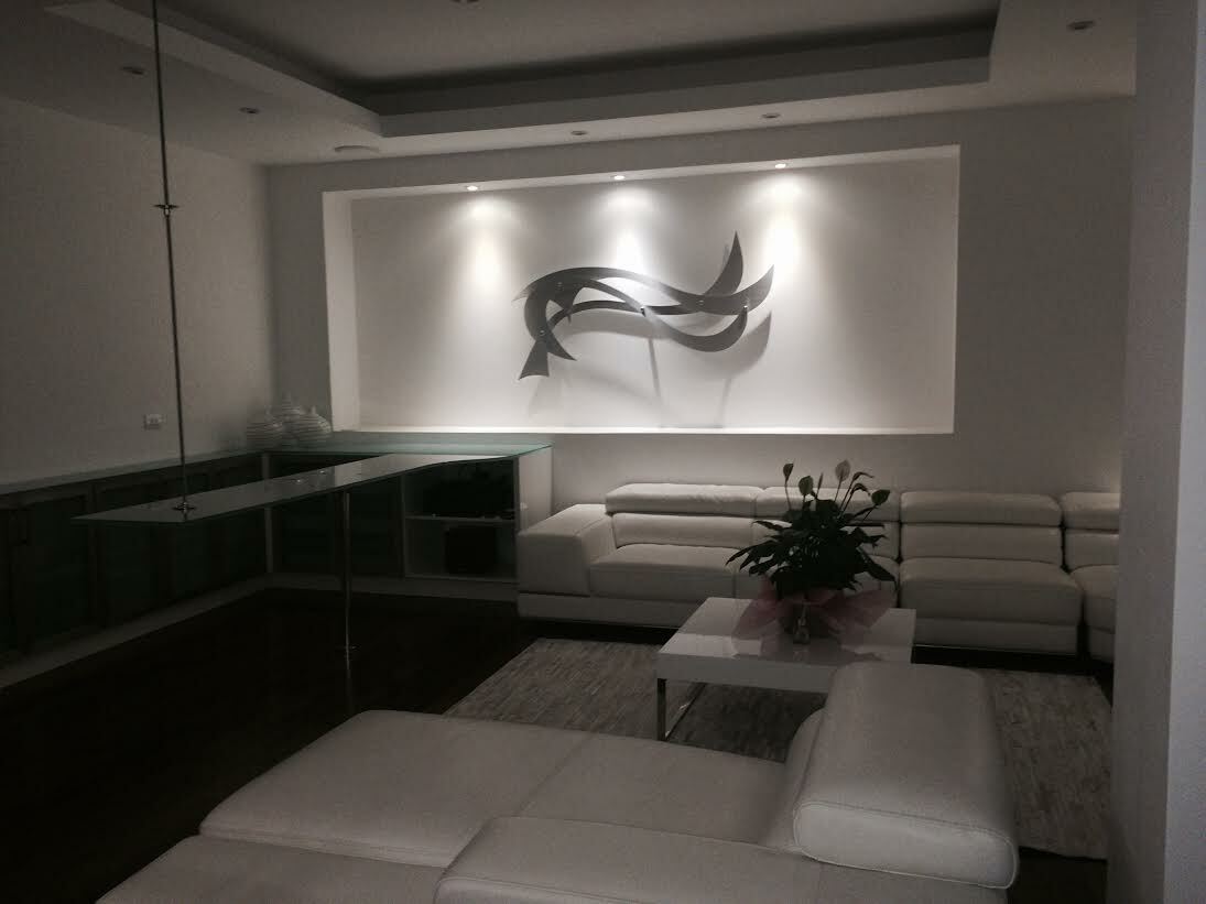 A modern living room with a sleek, minimalist design, featuring a custom metal wall art piece as the focal point. The artwork, composed of abstract, flowing shapes, is illuminated by subtle recessed lighting, creating soft shadows on the wall. The room is furnished with white leather sofas arranged in an L-shape, a glass coffee table with a potted plant, and clean, contemporary lines throughout the space. The overall ambiance is calm and sophisticated, emphasizing the elegance of the metal art installation.