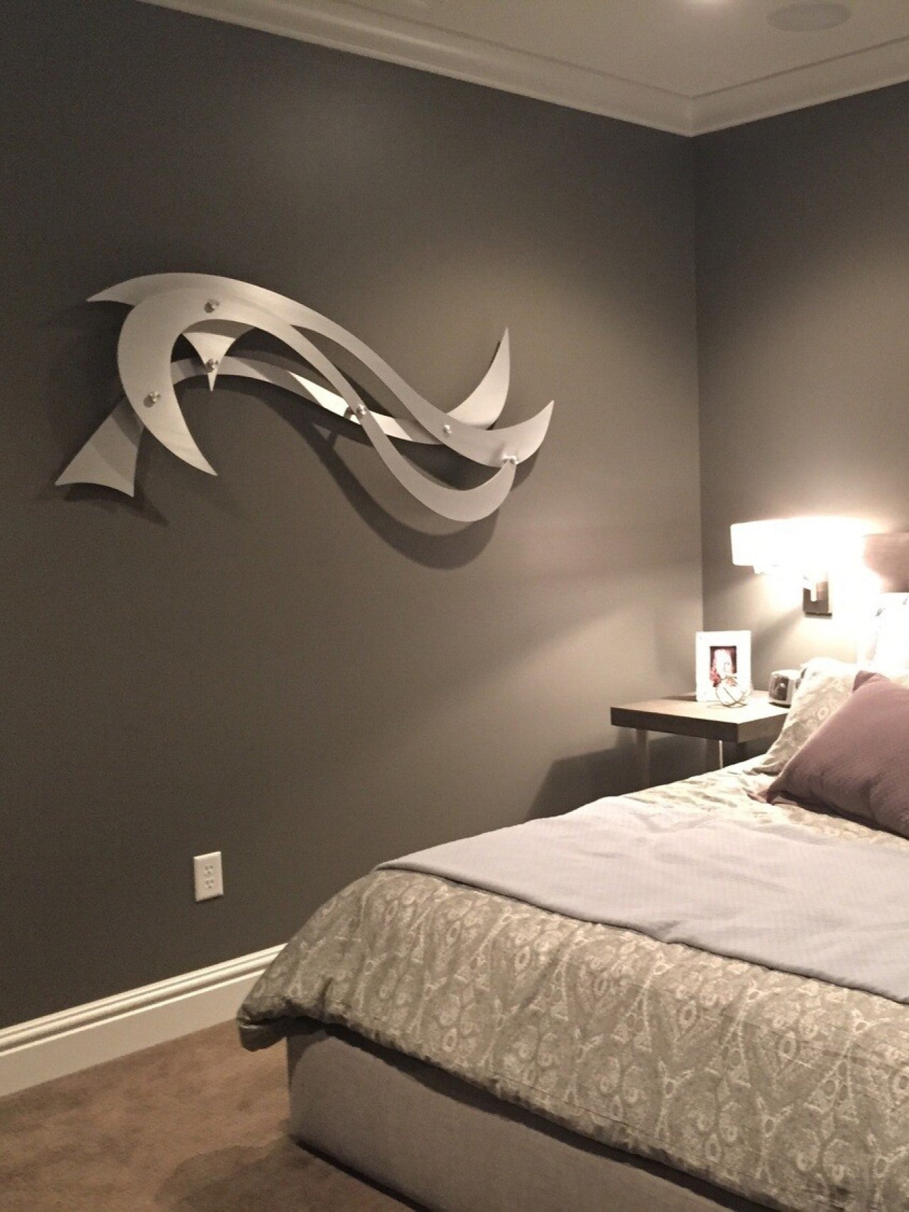 A sleek, modern metal wall sculpture with curved, wave-like shapes decorates a dark gray bedroom wall.